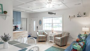 A bright, well-lit living room with a ceiling fan, a dining area with a table and chairs, and a kitchen area with blue cabinets and a coffee maker. - Bogue Bungalow: Oceanside Duplex, downtown Emerald Isle! (Emerald Isle)