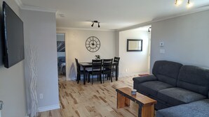 A living room with a grey sofa, wooden flooring, a dining table with chairs, a wall clock, and a ceiling fan. - Appartement Style Urbain Très Tranquille Situés a 5 min du Vieux Terrebonne (Laval)