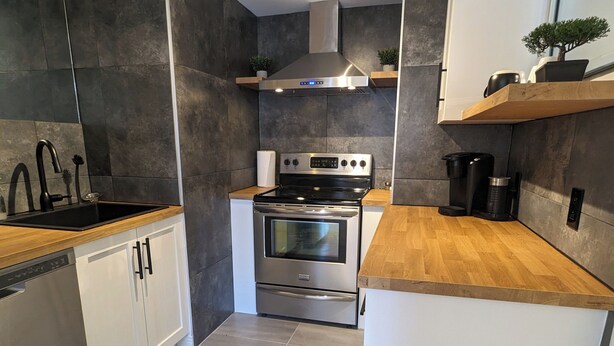 A modern kitchen with dark tiles, stainless steel appliances, and wooden countertops. - Appartement Style Urbain Très Tranquille Situés a 5 min du Vieux Terrebonne (Laval)