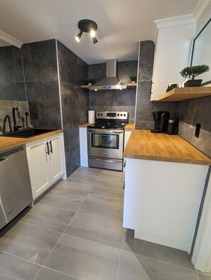 A modern kitchen with dark tiles, stainless steel appliances, and wooden countertops. - Appartement Style Urbain Très Tranquille Situés a 5 min du Vieux Terrebonne (Laval)