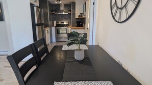 A modern kitchen with stainless steel appliances, a dining area with a black table and chairs, and a large wall clock. - Appartement Style Urbain Très Tranquille Situés a 5 min du Vieux Terrebonne (Laval)
