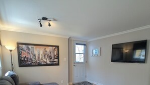 A living room with a sectional sofa, a coffee table, a painting, a door, a television, and a floor lamp. - Appartement Style Urbain Très Tranquille Situés a 5 min du Vieux Terrebonne (Laval)