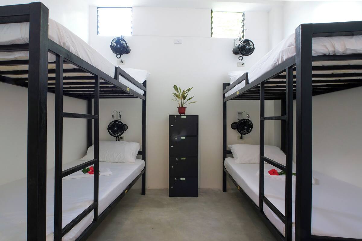 A dormitory room with bunk beds, a central cabinet, and wall-mounted fans.