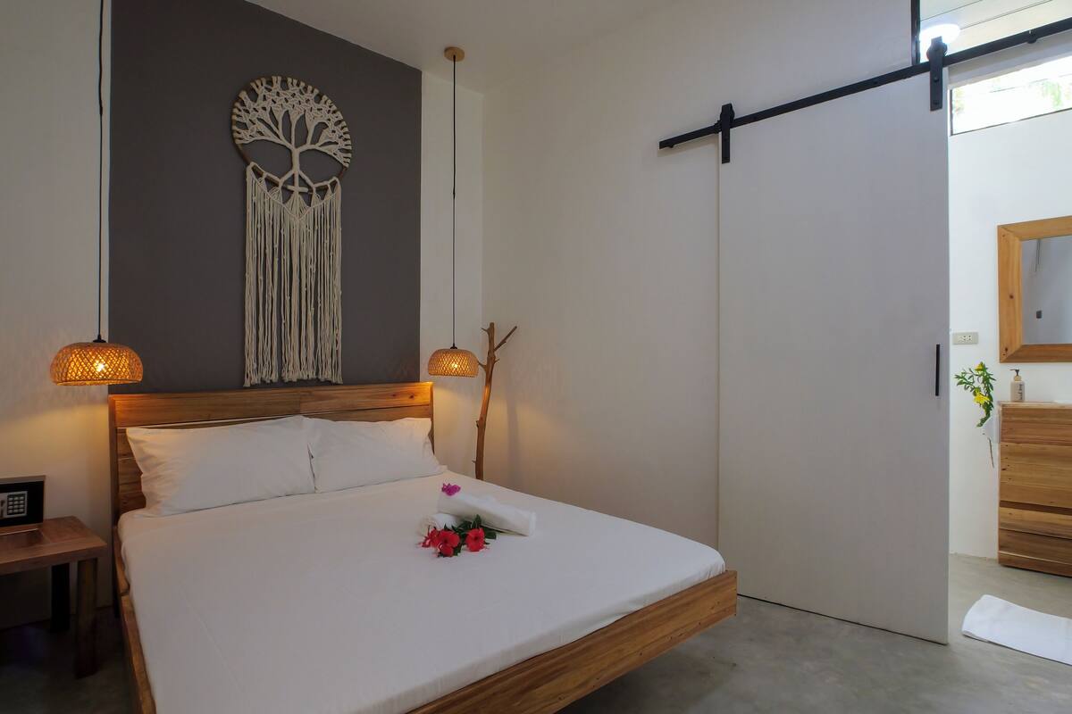 A bedroom with a bed, bedside lamps, a wall-mounted macrame tree, a wooden dresser, and a sliding door.