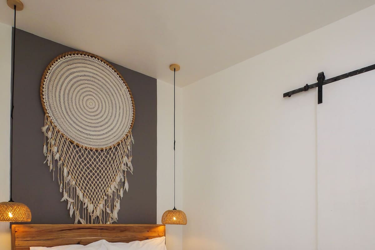 A modern bedroom with a wooden bed, white linens, a macramé wall hanging, and two pendant lights.