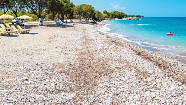 Una playa de guijarros con sillas de playa y sombrillas, un cielo azul despejado y mar tranquilo.