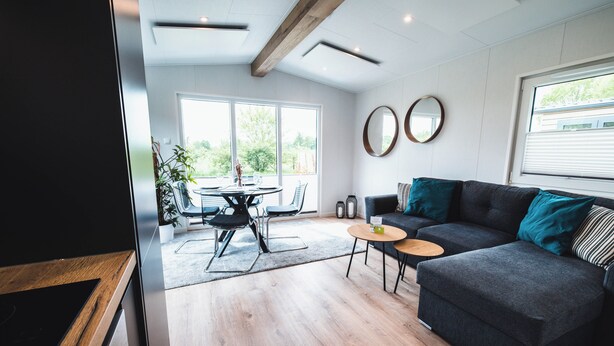 Een moderne woonkamer met een donkergrijze bank, een ronde salontafel en een eethoek met een tafel en stoelen. Twee ronde spiegels hangen aan de muur en een groot raam zorgt voor natuurlijk licht.