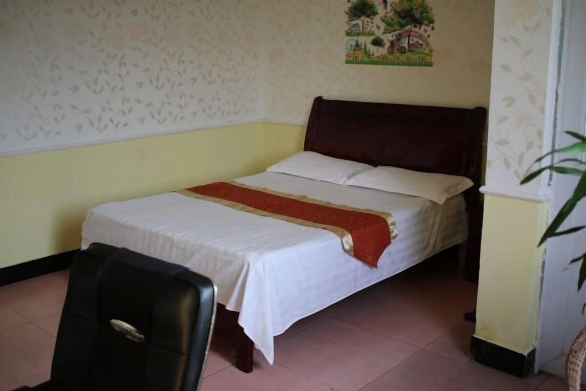 A single bed with a wooden headboard, a white bedspread, and a red runner. A black chair is positioned in front of the bed.