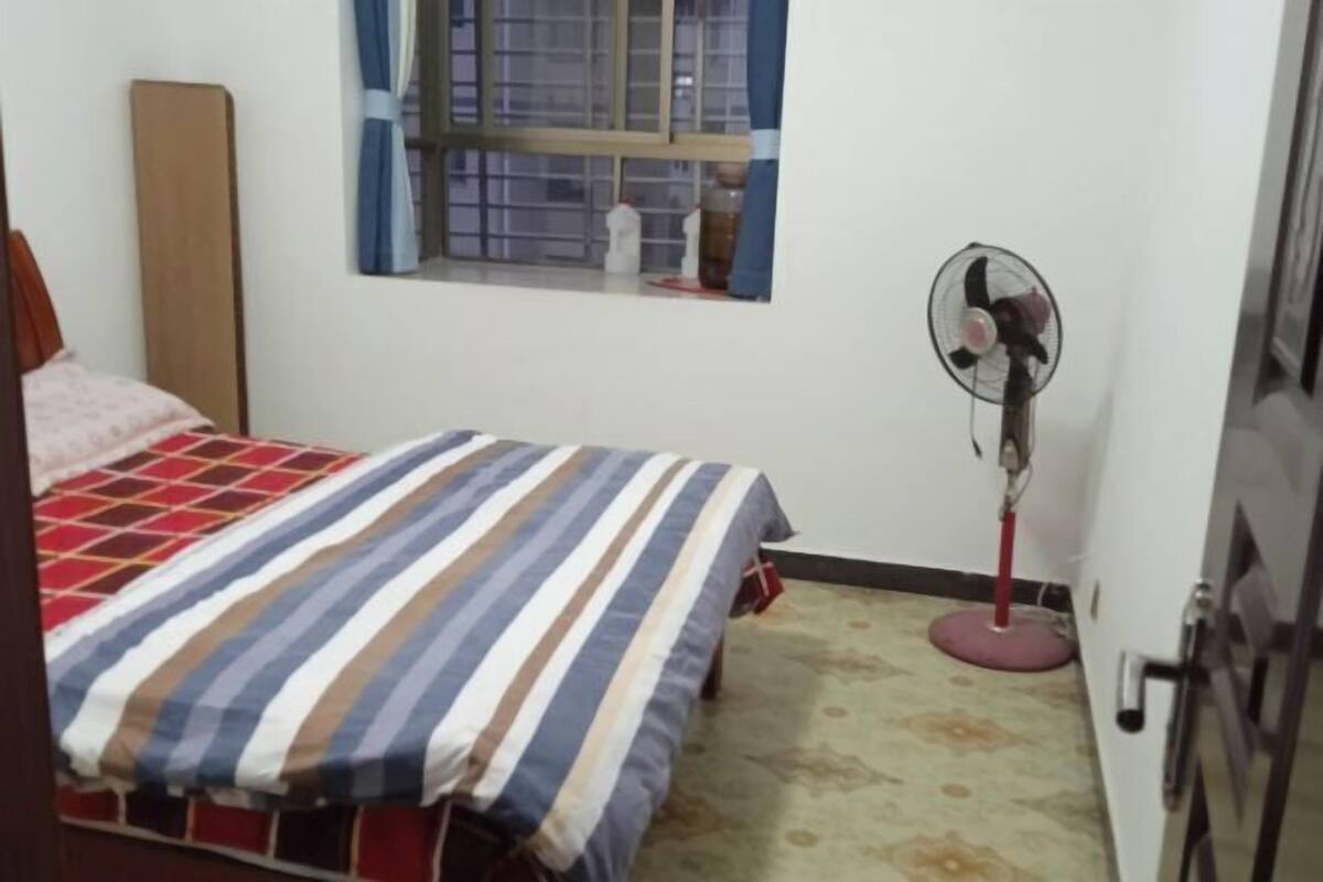 A bedroom with a bed, a standing fan, a window with curtains, and a door.