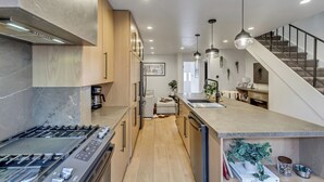 Una cocina moderna con una isla central, electrodomésticos de acero inoxidable, armarios de madera y una escalera que sube a un nivel superior.