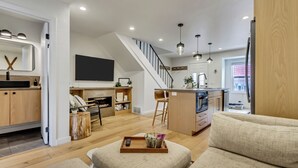 Una sala de estar moderna con un sofá, una mesa de centro con una bandeja, y una zona de cocina con un televisor, armarios de madera y una escalera.