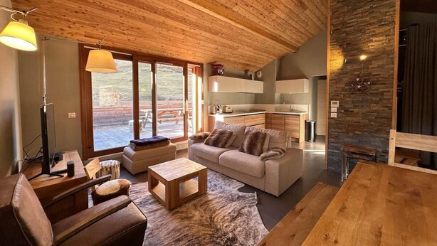 A spacious living area with a wooden ceiling, a sofa, a coffee table, and a dining table. There is a kitchen area with a stone wall and a large window. - 5th floor, balcony, tv, ski locker, 65m², Vars (Vars)