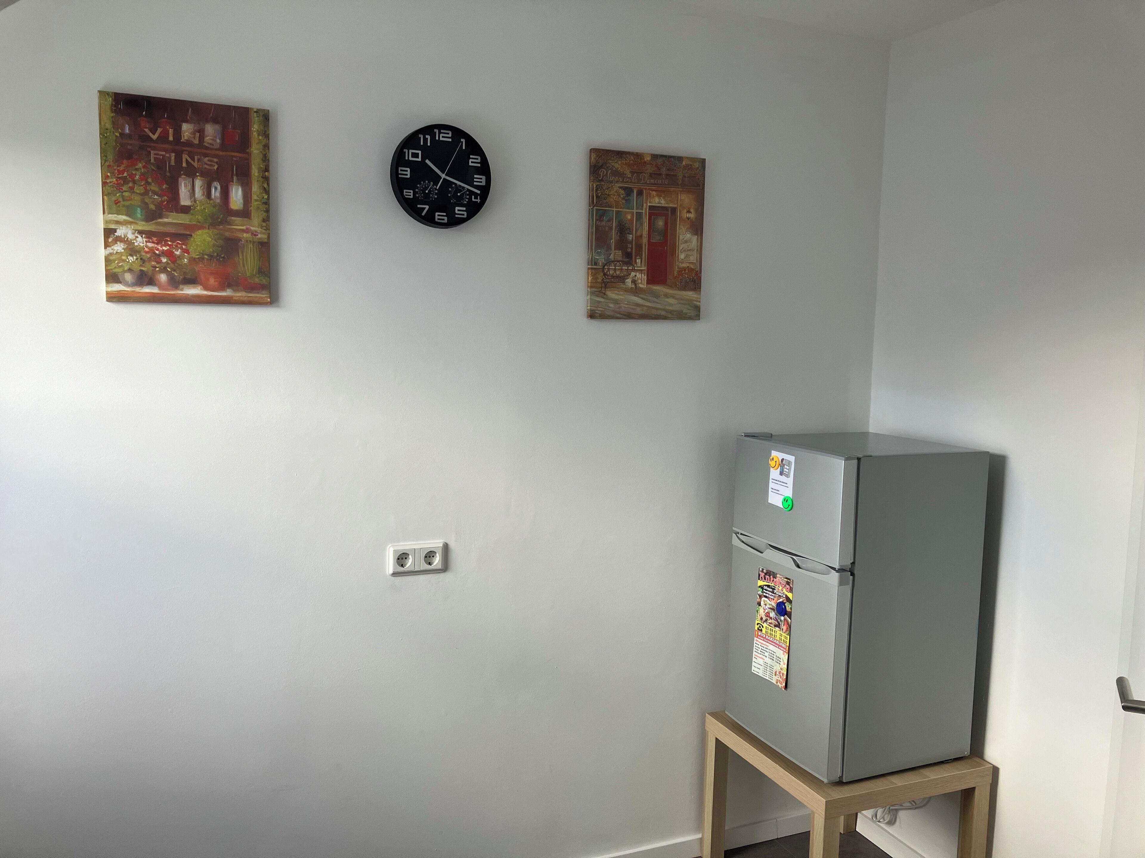 Una habitación con un refrigerador sobre un soporte de madera, un reloj de pared y dos cuadros.