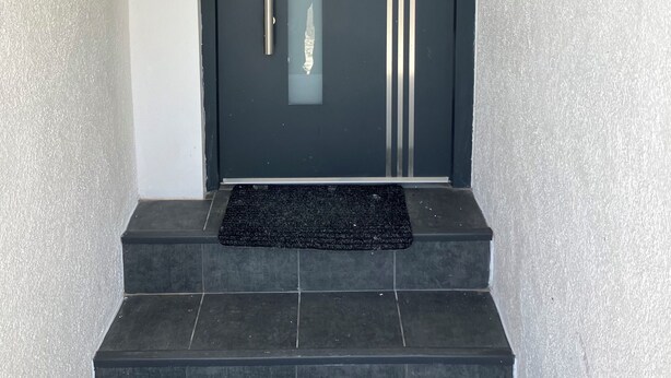 A black door with a vertical handle and a peephole, flanked by two vertical stripes, and a black mat at the base of the door. - Borsch Holiday Apartment 2 (Geisa)