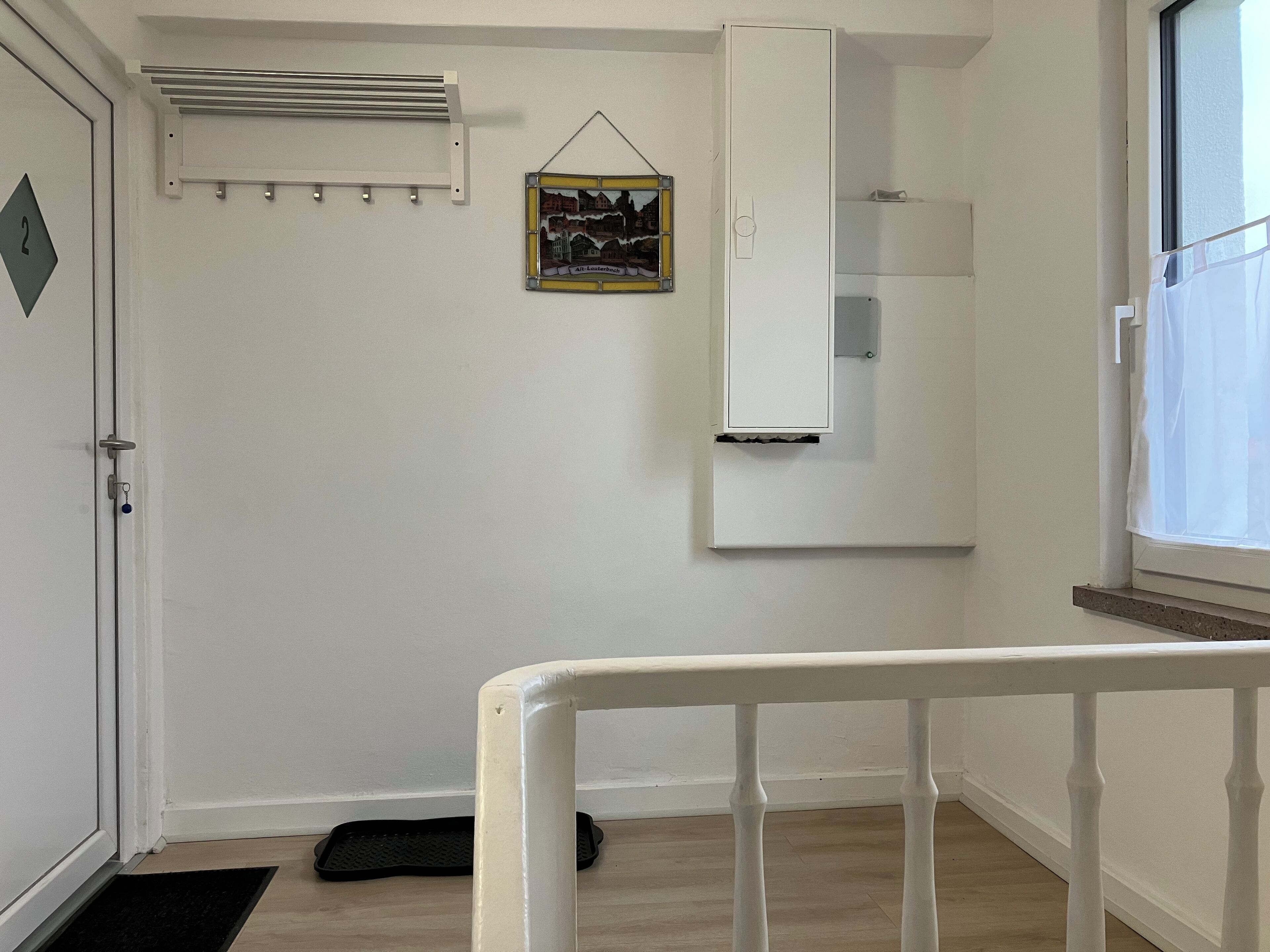 Et værelse med en hvid dør, en radiator, et hvidt skab, et billede i ramme og et vindue med gardin.