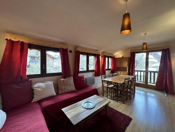 A cozy living room with a sofa, a dining table, and chairs. There are large windows with curtains, a wooden ceiling, and a hanging light fixture.