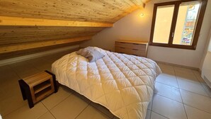 A bedroom with a wooden ceiling, a bed with a white duvet, a small wooden stool, a nightstand, and a window with a view. - 3rd floor, balcony, tv, ski locker, 80m², Vars (Vars)
