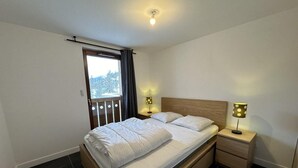 Une chambre à coucher avec un lit, deux tables de chevet, une fenêtre avec des rideaux et un luminaire au plafond.