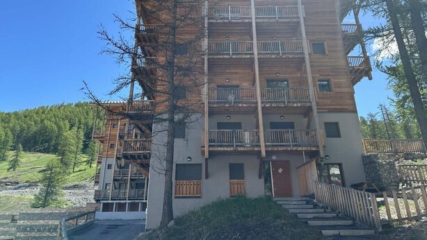 Un immeuble en bois à plusieurs étages, doté de balcons, d’une entrée en pierre et offrant une vue dégagée sur la forêt environnante.