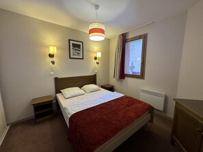A bedroom with a bed, bedside lamps, a window with curtains, a radiator, and a wooden nightstand.
