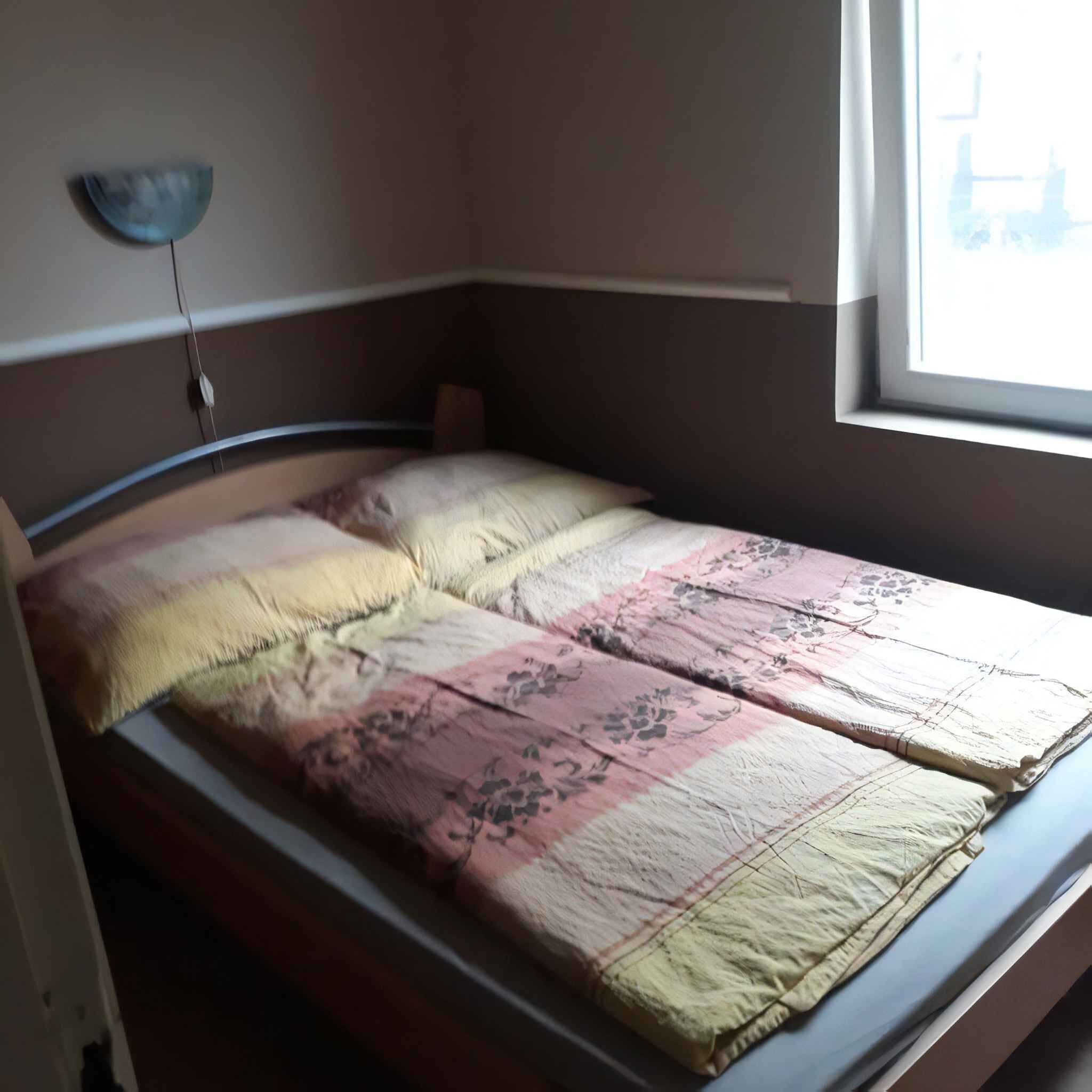 Sänky, jossa on kukkakuviollinen päiväpeite ja koristetyynyjä, yöpöytä ja seinään kiinnitetty valaisin.