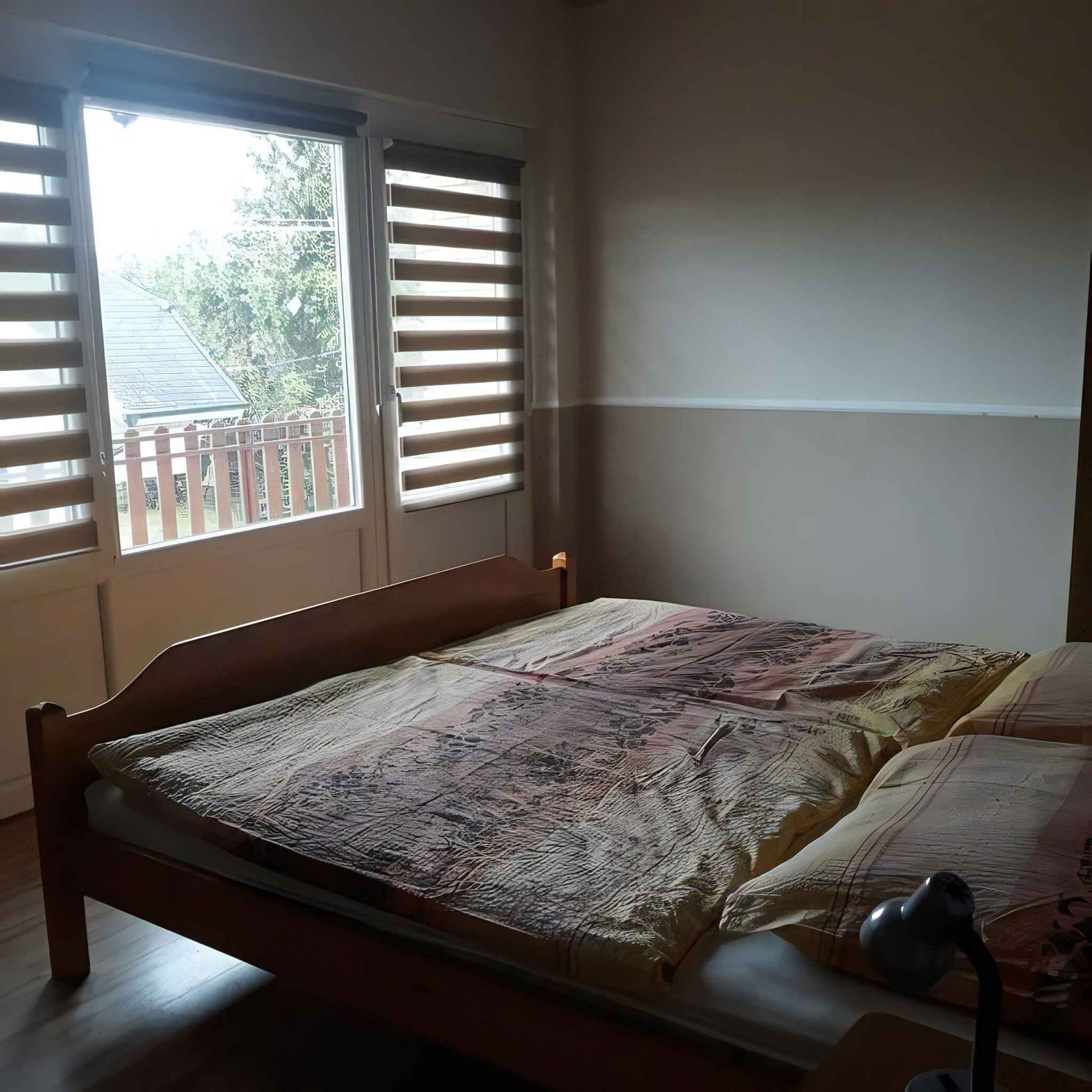 Une chambre à coucher avec un lit en bois, une fenêtre avec des stores et une vue sur une clôture et des arbres à l’extérieur.