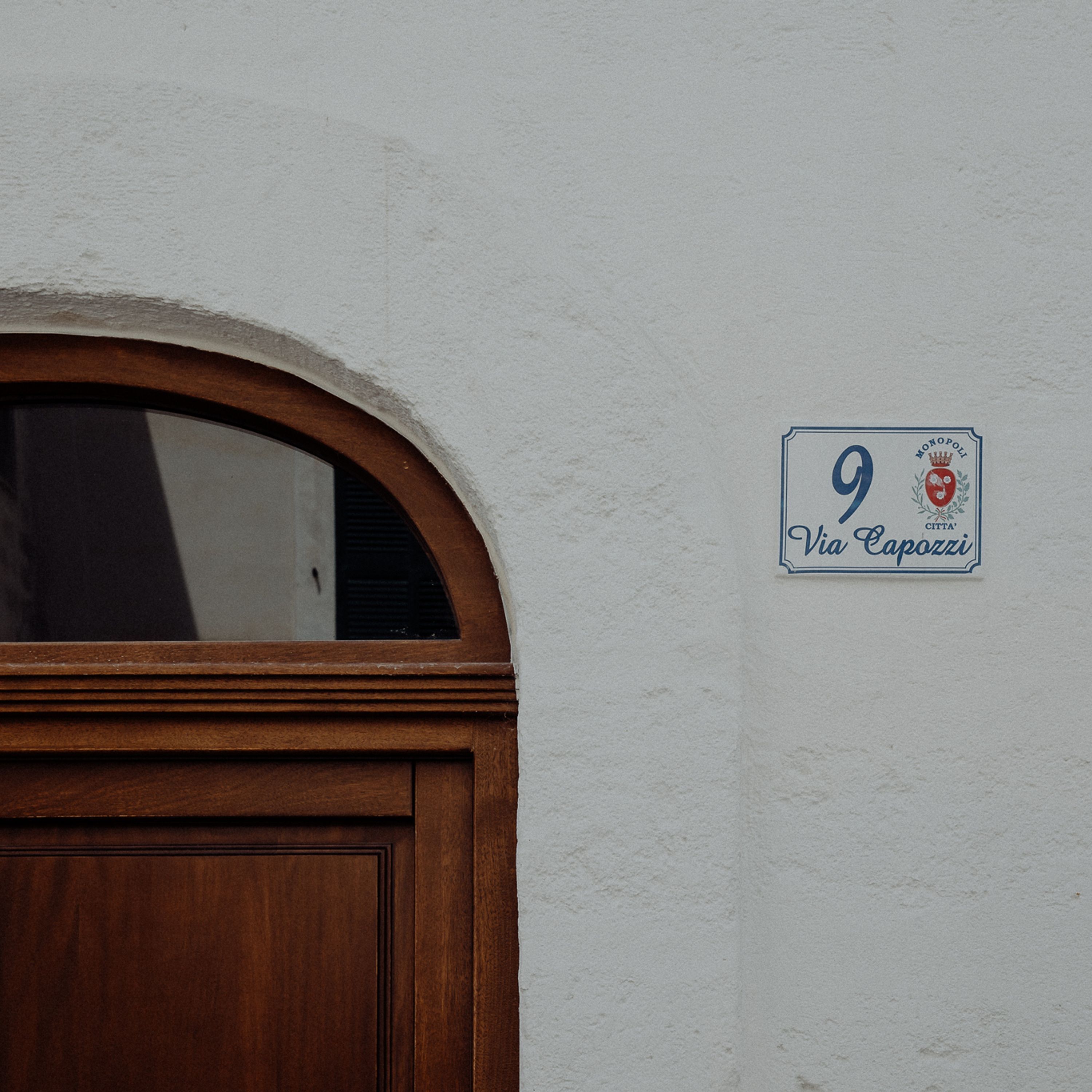 Entrada de un edificio con puerta de madera y pared blanca. Se ve un cartel con el número 9 y el texto 'Via Capozzi'.