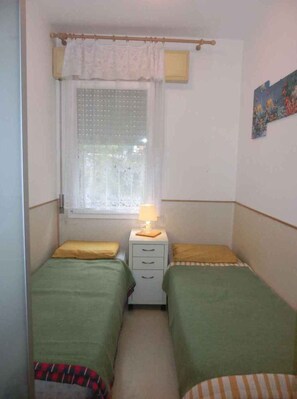 A small, narrow room with two single beds, a window with a curtain, a bedside table with a lamp, and a small piece of wall art.