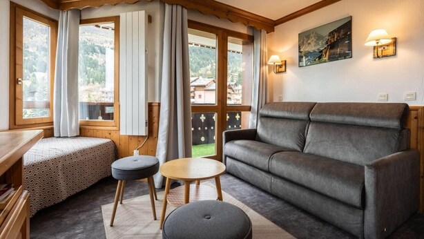 Una habitación acogedora con un sofá, dos reposapiés, una mesita redonda y un banco de madera. Amplias ventanas ofrecen vistas del exterior.