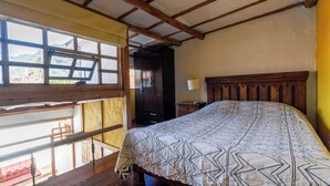 Une chambre avec un plafond en bois, un grand lit recouvert d’une couvre-lit à motifs, une table de chevet avec une lampe et une fenêtre donnant sur un paysage verdoyant.