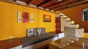 Un salon avec un plafond en bois, un canapé avec des coussins, une cheminée et deux tableaux accrochés au mur.