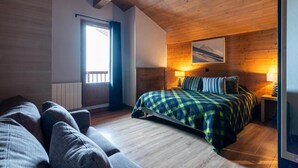 A cozy bedroom with a wooden ceiling, a bed with a checkered blanket, a nightstand with a lamp, and a window with curtains.