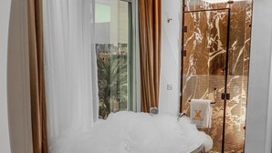 Une salle de bain moderne avec une grande baignoire remplie de mousse, une douche en verre et une vue sur les palmiers par la fenêtre.