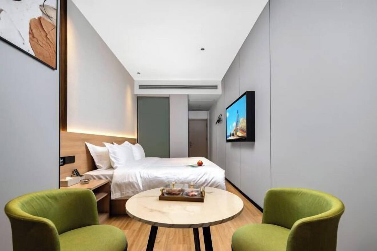 Una habitación de hotel moderna con una cama, un televisor montado en la pared y dos sillas verdes alrededor de una mesita redonda.