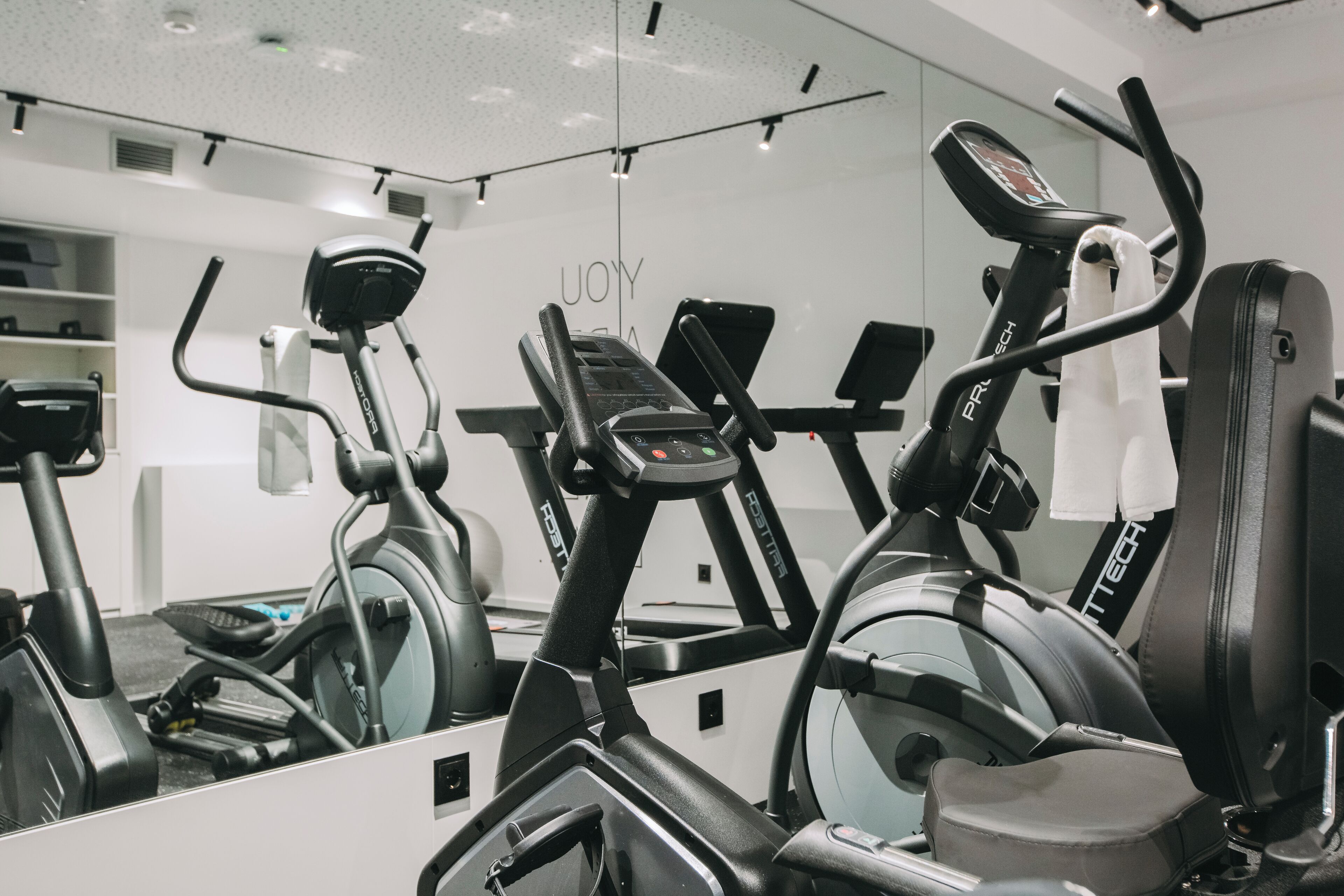 A gym with various exercise machines, including a treadmill and an elliptical, with a mirrored wall reflecting the equipment.