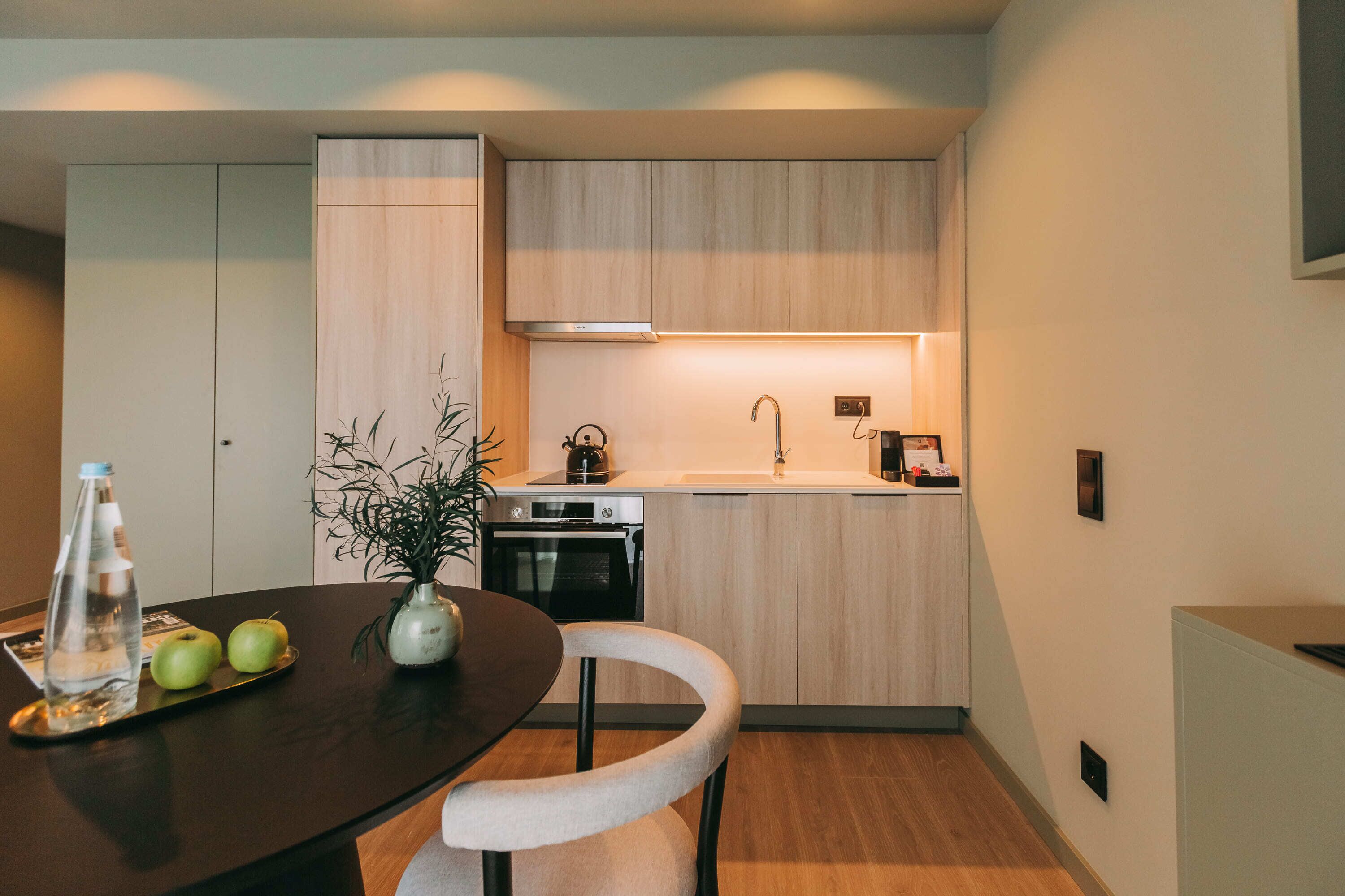 A modern kitchen with wooden cabinets, a built-in oven, and a sink. There is a dining table with a vase of plants and a bottle of water.