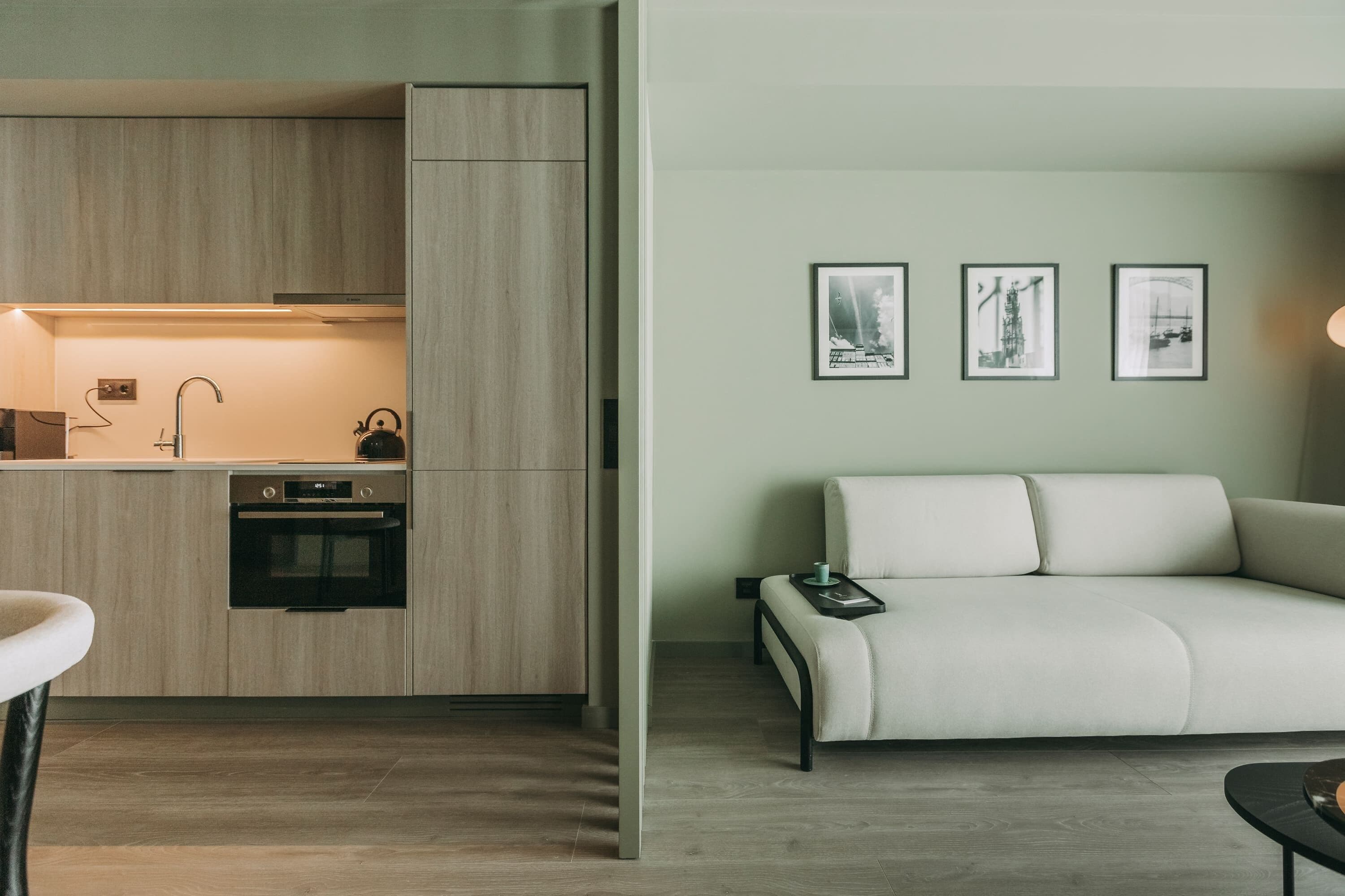 Una cocina moderna con armarios de madera, un horno empotrado y un lavamanos. Hay un sofá blanco y una mesita con una taza encima. Tres cuadros enmarcados están colgados en la pared.