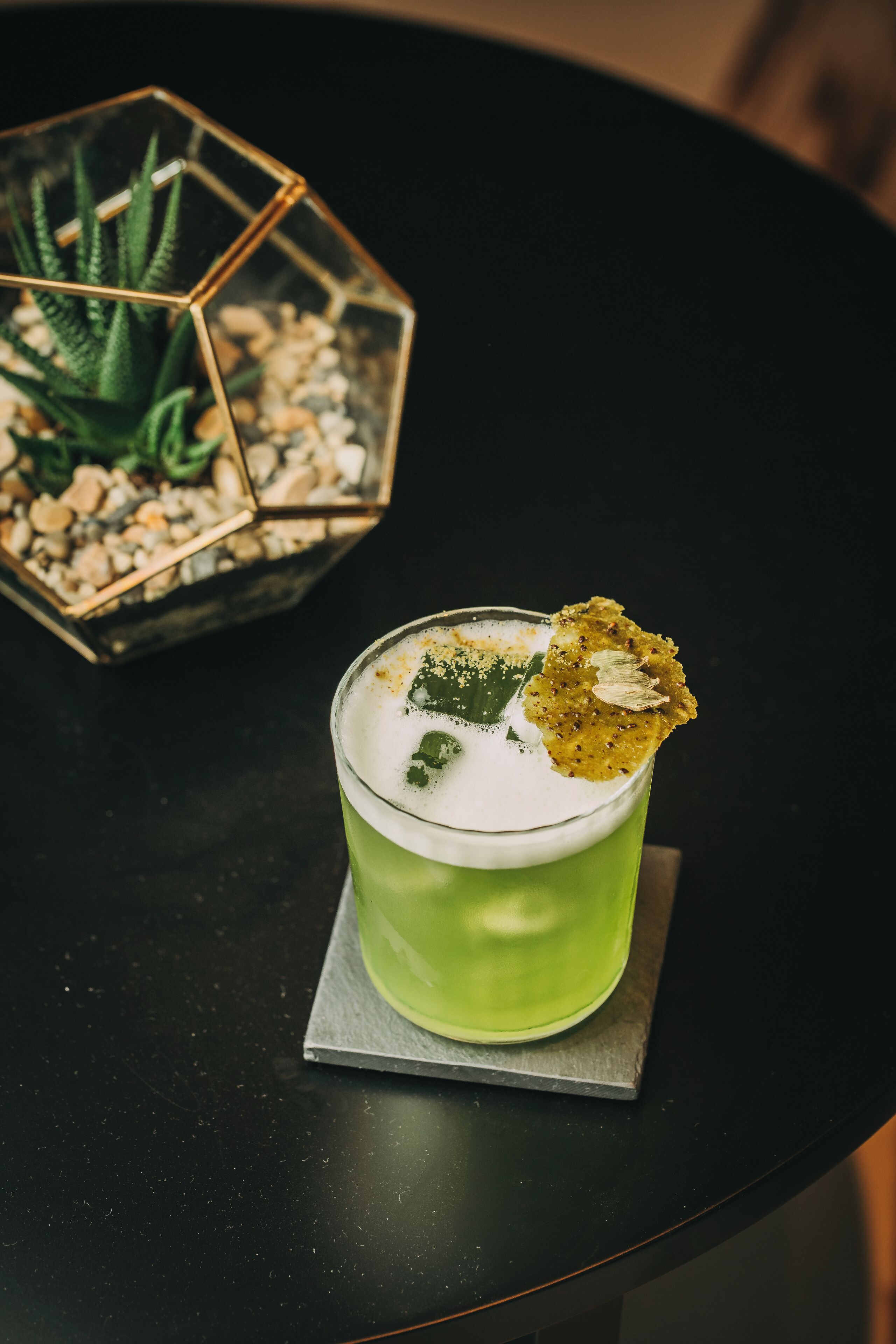 A cocktail with a garnish on a coaster, placed on a table with a geometric glass container and a succulent plant in the background.