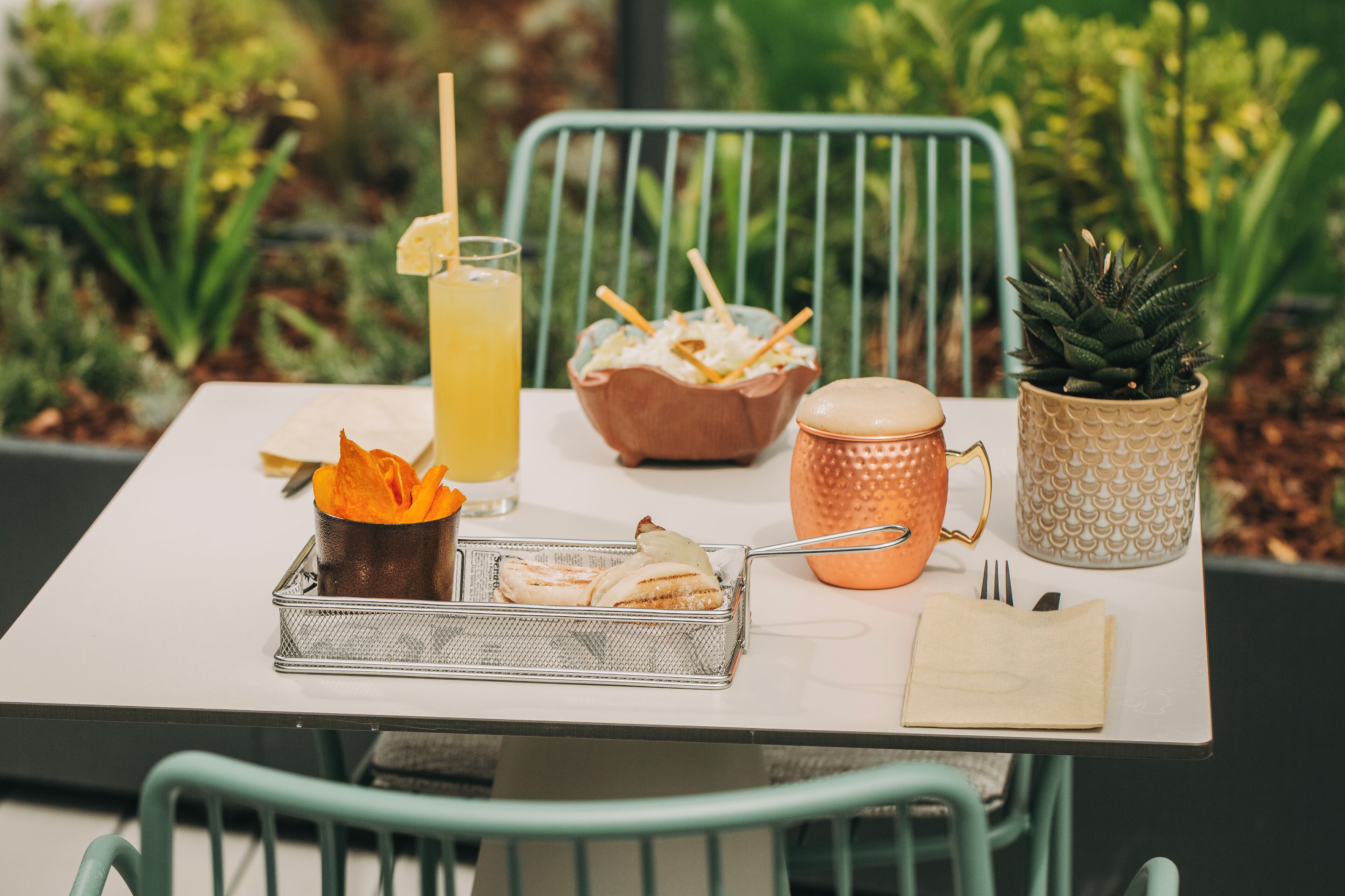 A table with a metal basket containing food items, a glass of a yellow beverage, a bowl of snacks, a copper mug, and a small potted plant.