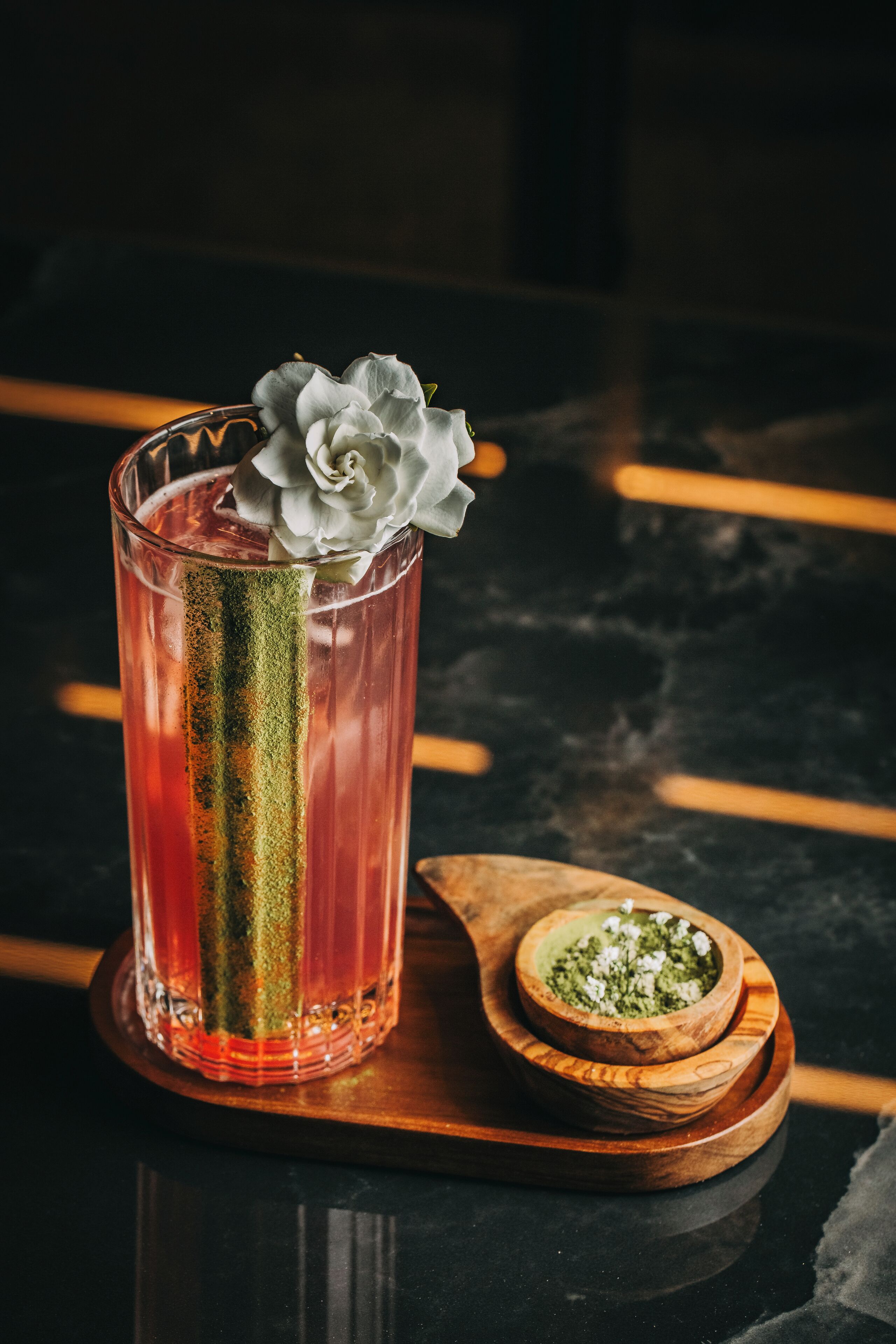 A tall glass with a rose garnish, a green stripe, and a wooden spoon with a green substance beside it.