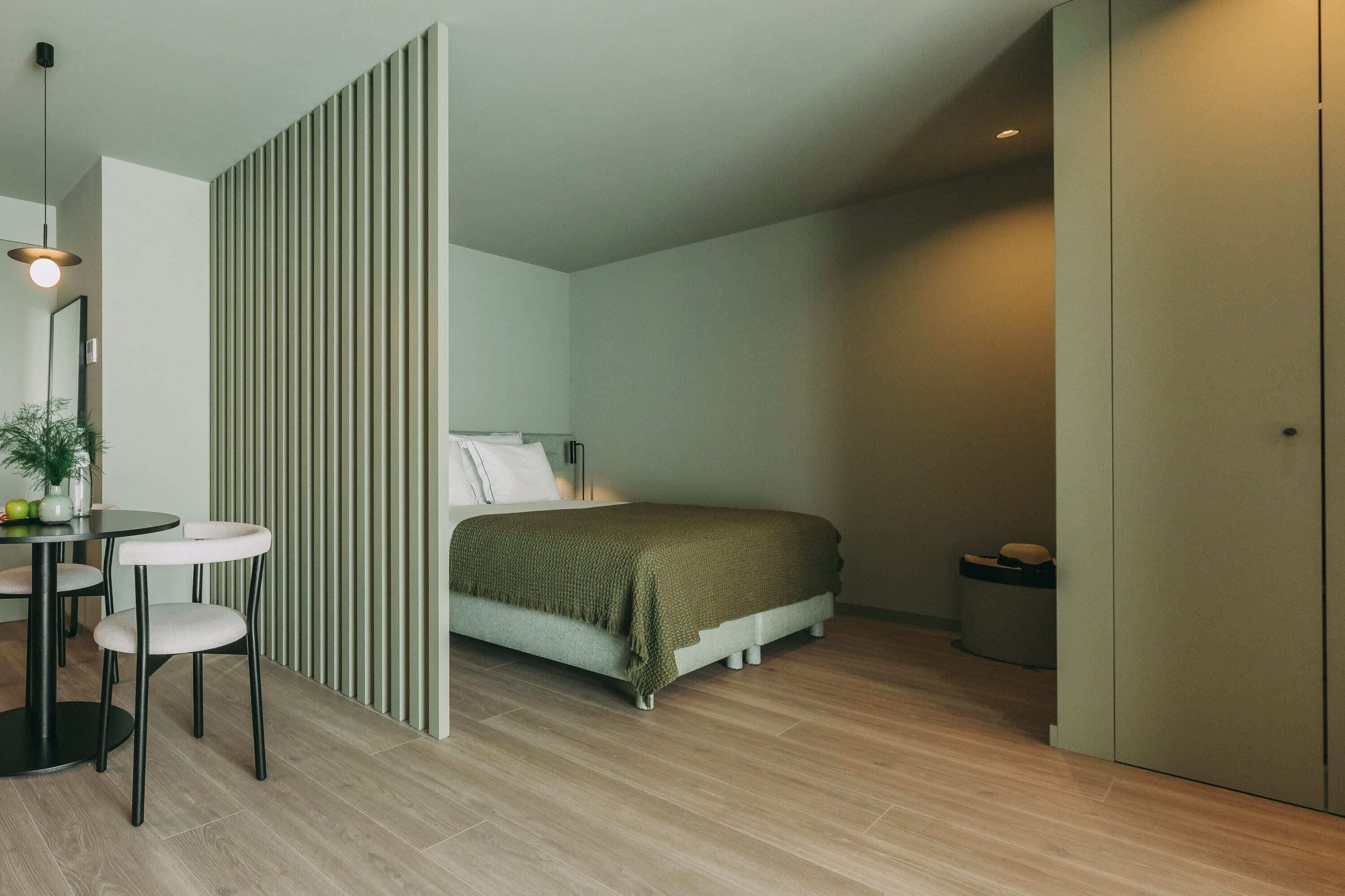 Un dormitorio con cama, mesita de noche y una silla. Se observa una pared divisoria con líneas verticales, una pequeña mesa redonda con una silla y una planta en maceta.