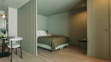 Un dormitorio con cama, mesita de noche y una silla. Se observa una pared divisoria con líneas verticales, una pequeña mesa redonda con una silla y una planta en maceta.