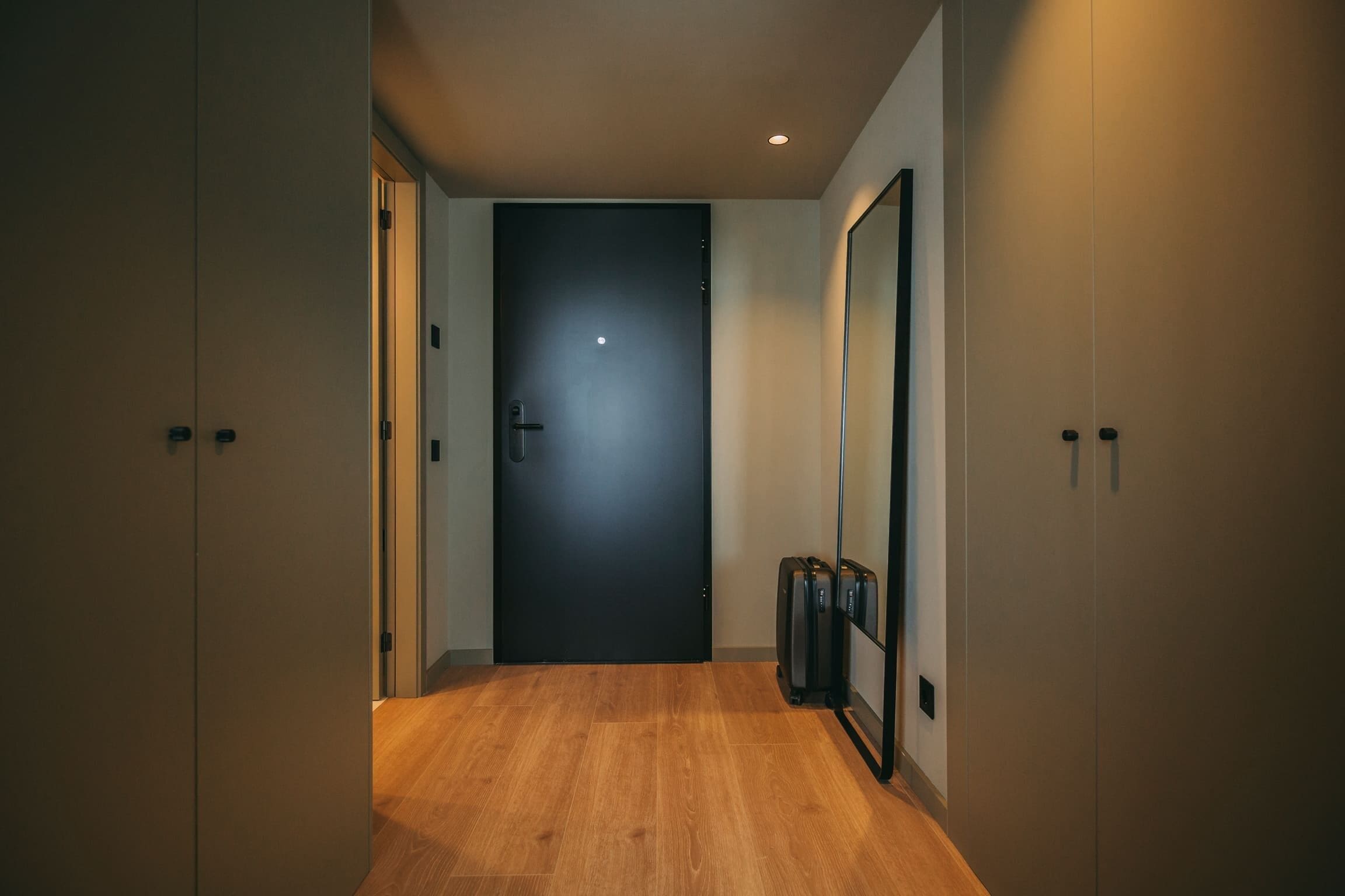 A hallway with a wooden floor, a black door, and a luggage rack against the wall.