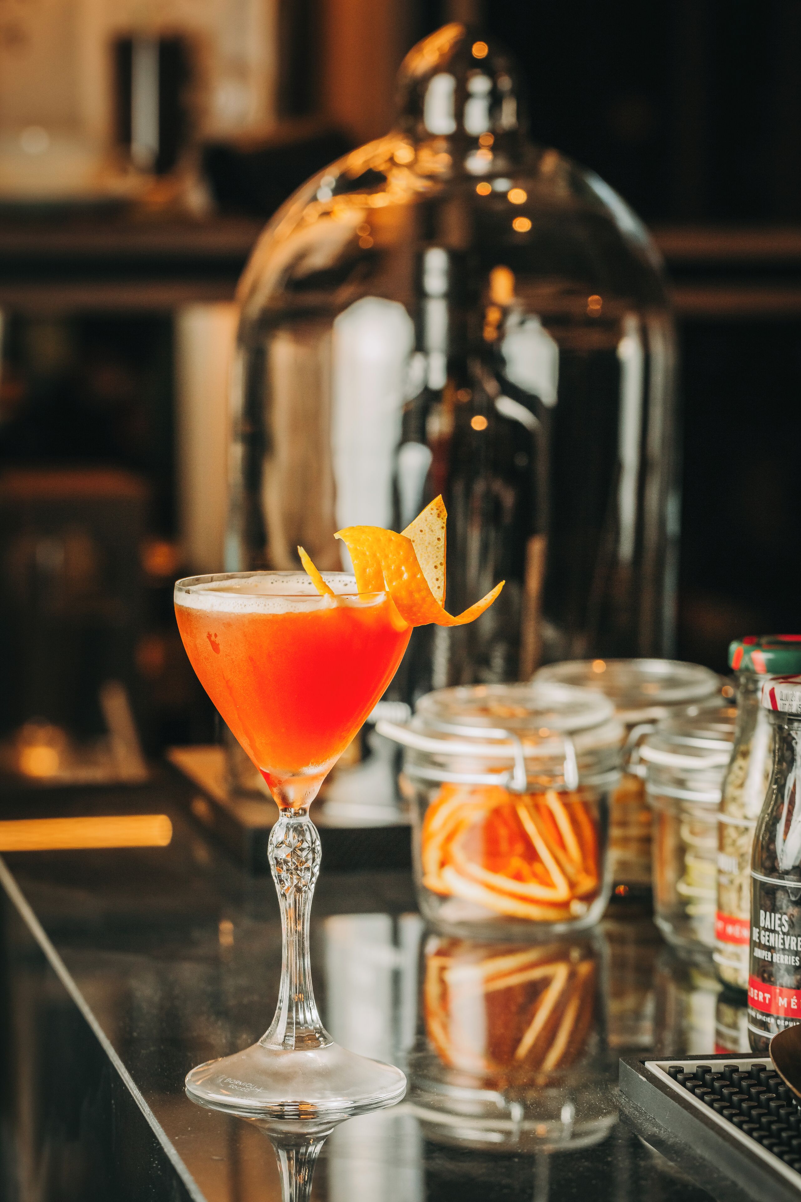 Un cocktail servi dans un verre à martini, décoré d’un zeste d’orange, avec un shaker et un bocal rempli de zestes d’orange sur le comptoir du bar.