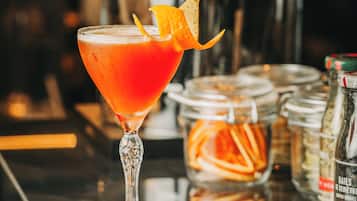 Un cocktail servi dans un verre à martini, décoré d’un zeste d’orange, avec un shaker et un bocal rempli de zestes d’orange sur le comptoir du bar.
