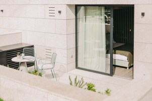 Un espace extérieur moderne aménagé avec une table et des chaises, une petite plante en pot et une porte vitrée donnant sur une chambre à coucher équipée d’un lit et de rideaux.