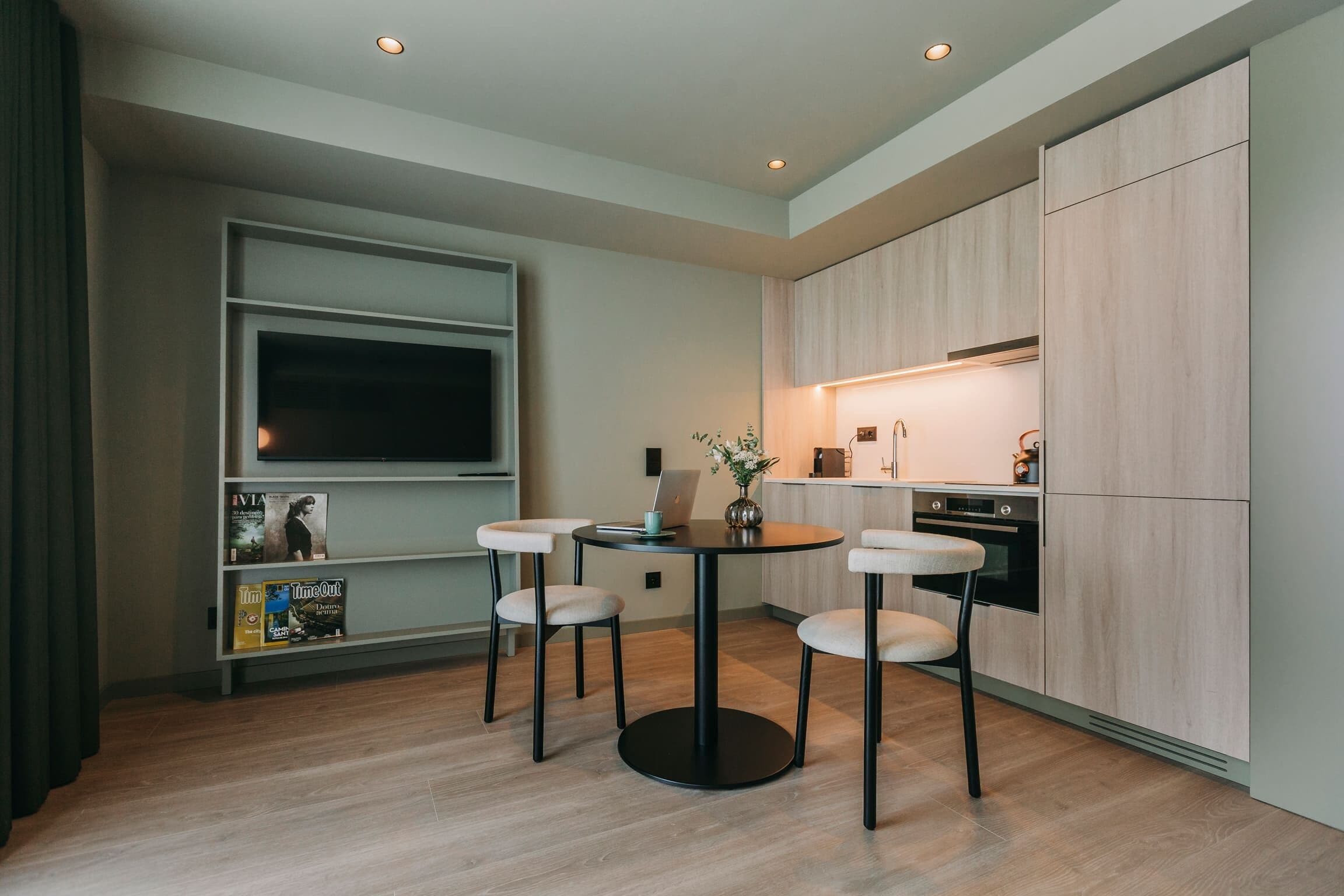 A modern kitchen with a built-in oven, a dining area with a round table and four chairs, and a large window with a TV.