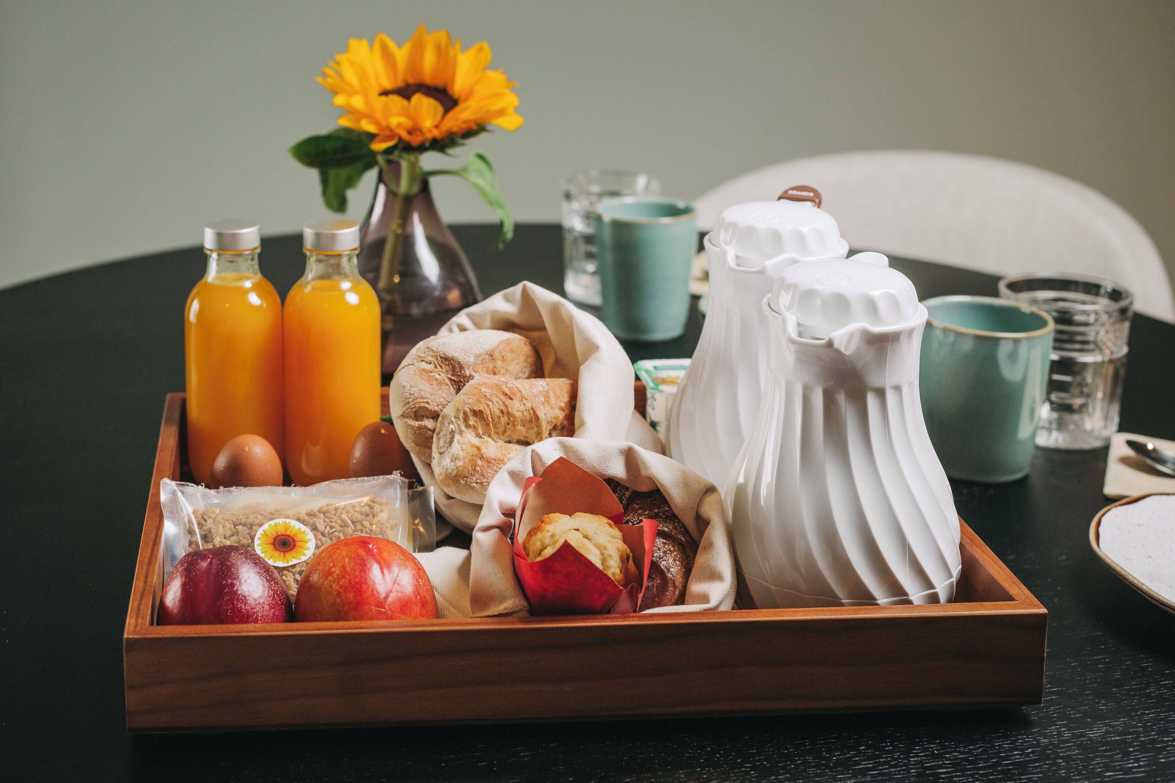 Een houten dienblad met ontbijtartikelen, waaronder brood, gebak, fruit en flesjes sap.