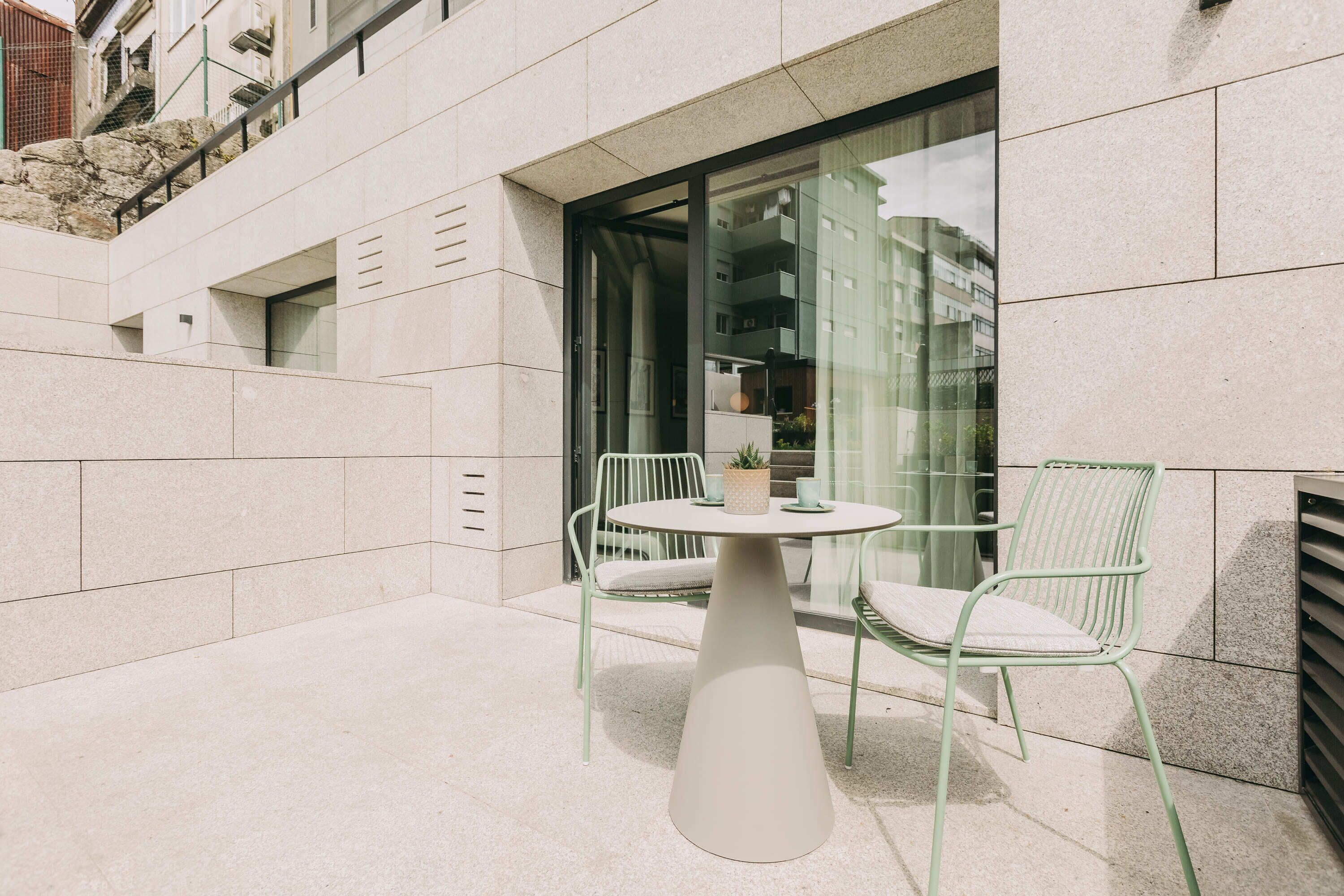 A modern outdoor seating area with a round table and two chairs, a potted plant, and a glass door leading to an interior space.
