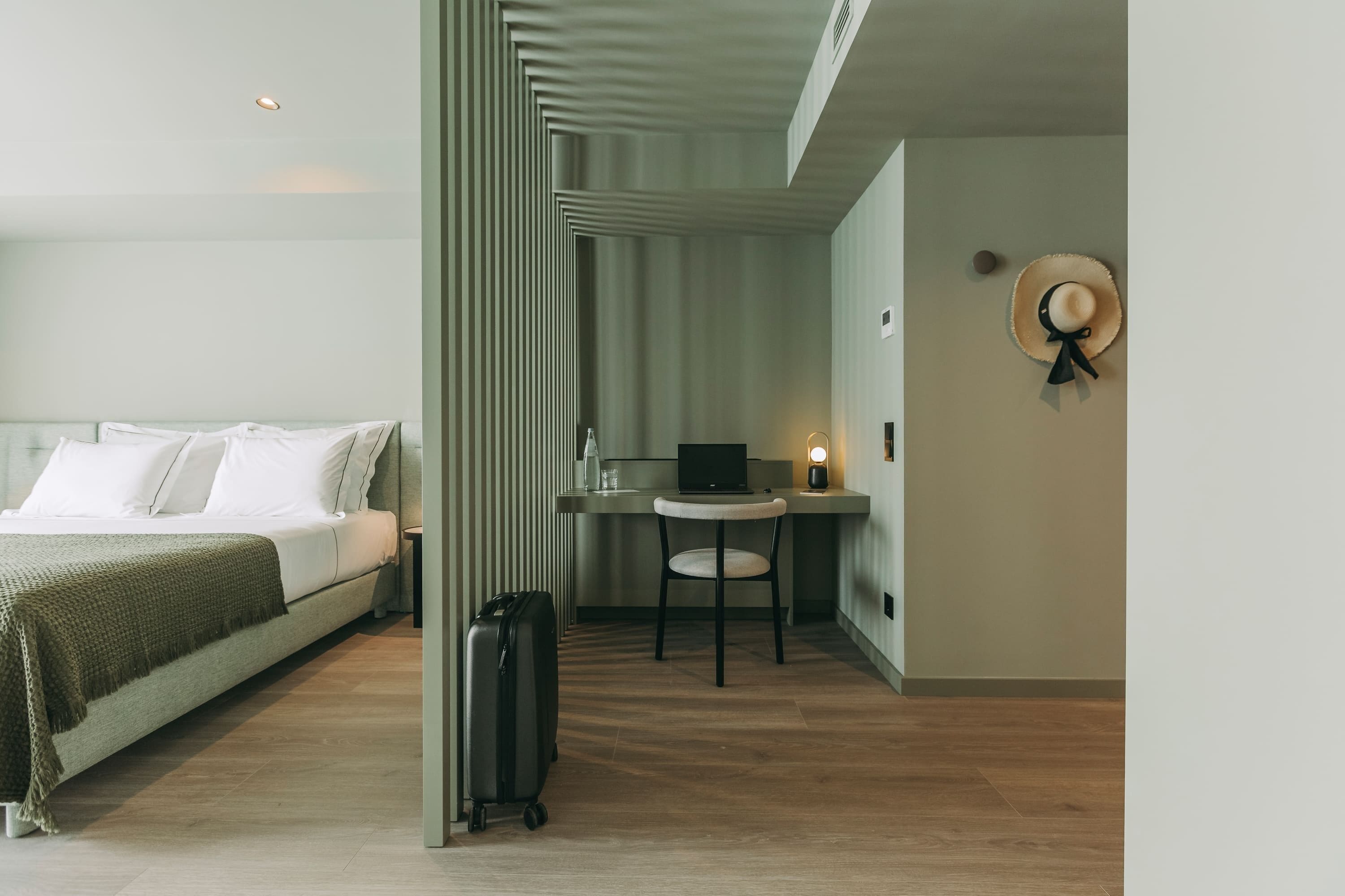 Ein modernes Hotelzimmer mit einem Bett, einem Schreibtisch, einem Laptop, einem Koffer und einem Hut an der Wand.